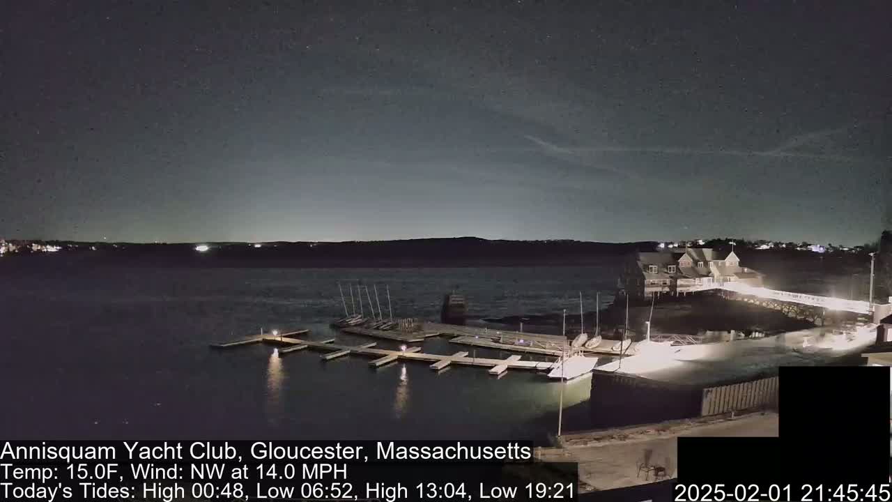 A nighttime view of a marina with docked sailboats under a partly cloudy sky, with a building lit up in the background.