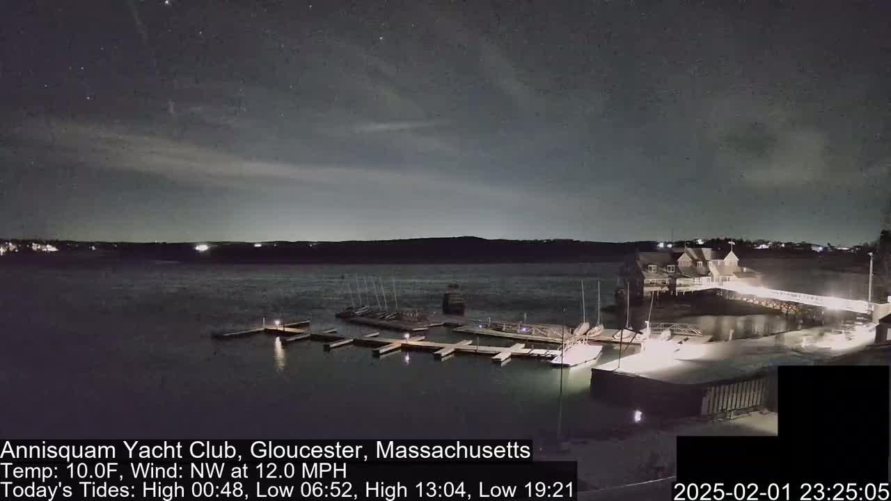 A nighttime view of a marina with boats docked, a clubhouse, and a calm body of water under a partly cloudy sky with stars visible.