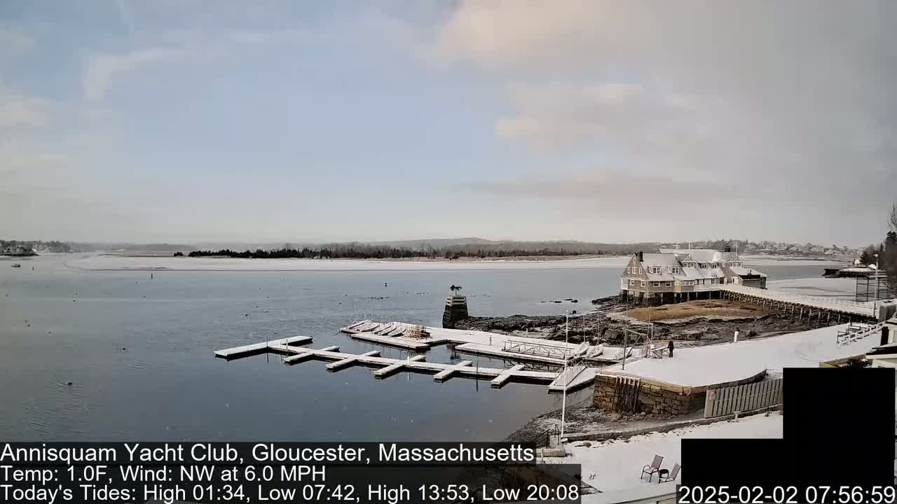 A snow-covered marina with a clubhouse and calm, partially frozen water under a mostly cloudy sky.