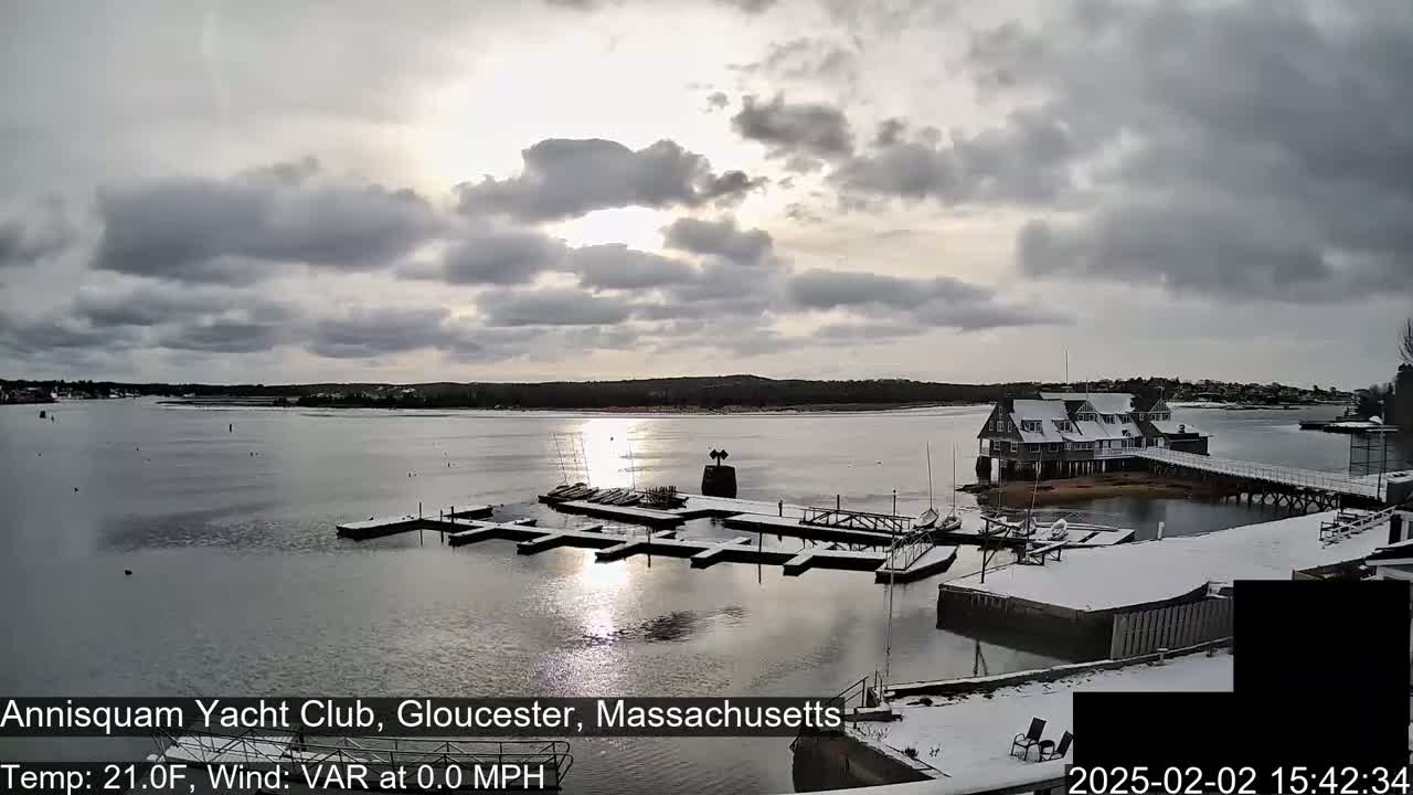 A snow-covered yacht club and marina sit on a calm body of water under a partly cloudy sky.