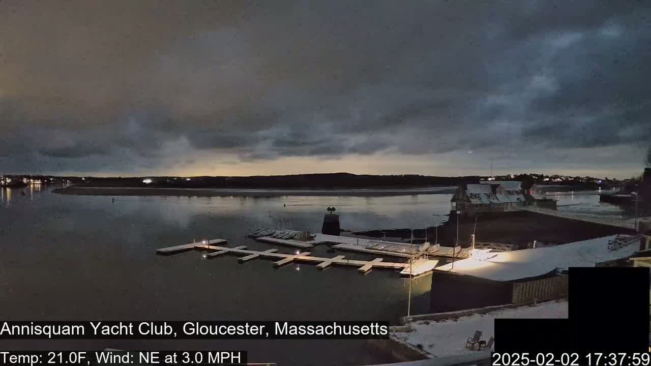A snow-dusted yacht club sits on a calm, dark body of water under a cloudy twilight sky.