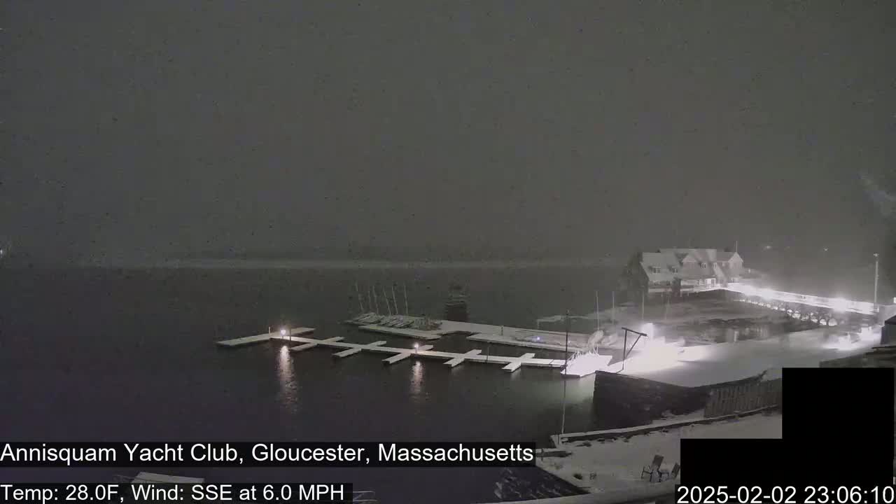 A snow-covered yacht club sits on a dark body of water during a light snowfall.
