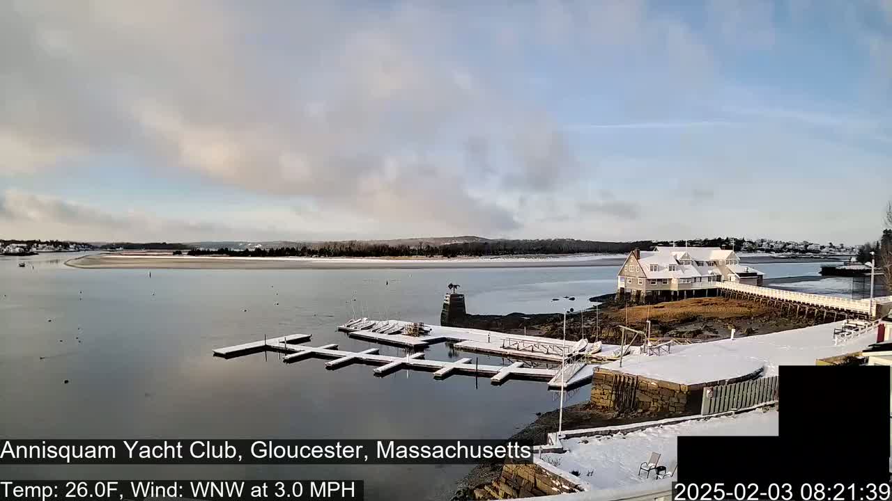 A snow-covered marina with docks and a building sits on a calm body of water under a mostly cloudy sky.