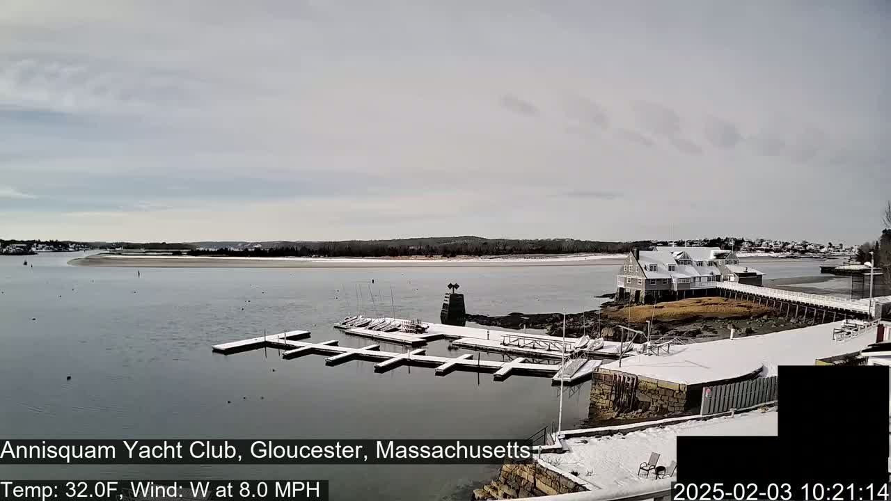 A snow-covered marina with docked boats sits on a calm body of water under a cloudy sky.