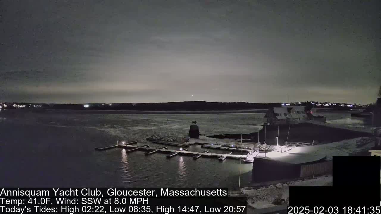 A nighttime view of a marina with docked boats, a building, and a calm body of water under a dark, cloudy sky.