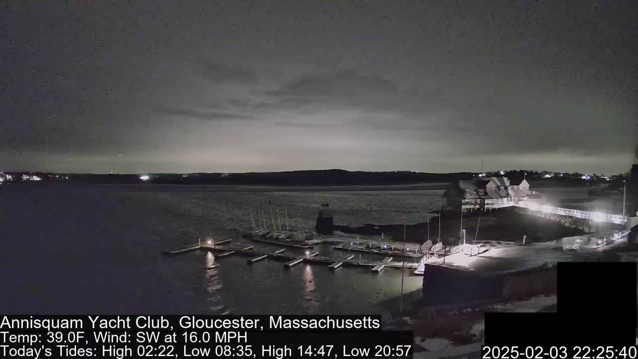 A nighttime view of a marina with docked boats, a clubhouse, and a snow-covered walkway under a dark, cloudy sky.