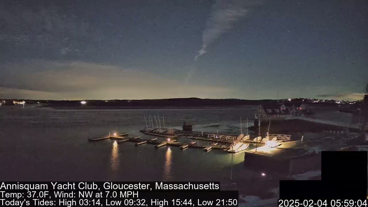 A nighttime view of a marina with boats docked, under a partly cloudy sky with a northwest wind.