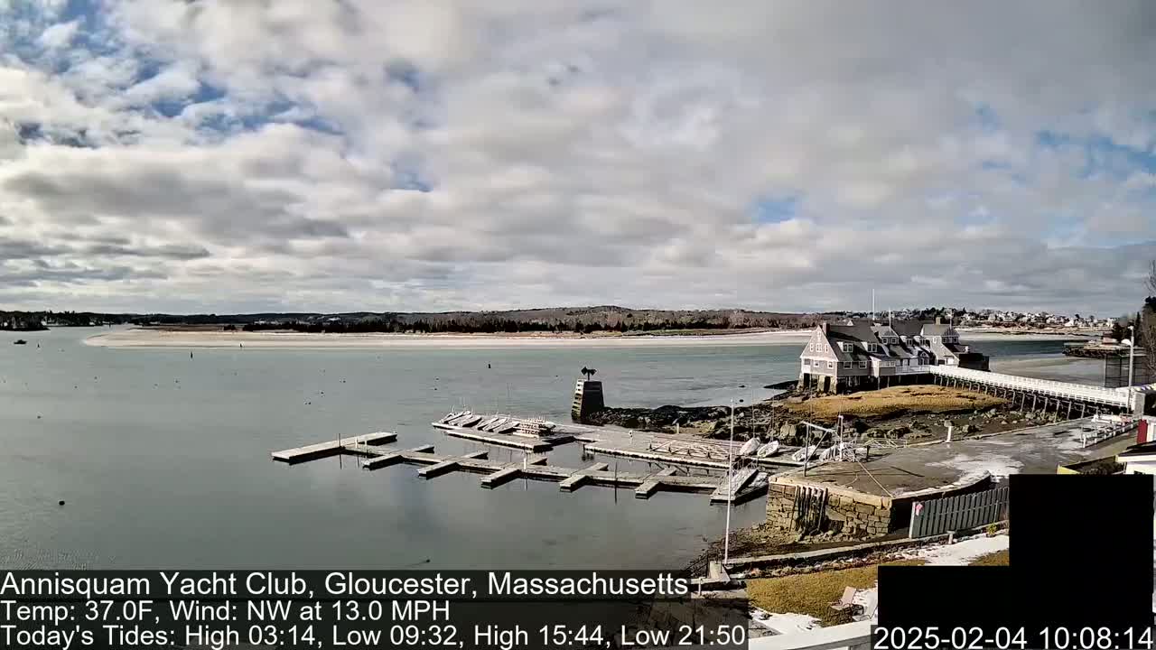 A partly cloudy day shows a marina with several boats docked, a clubhouse on a small island, and a wide sandy beach across the water.