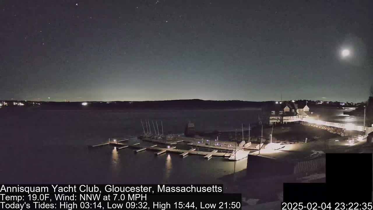 A moonlit night scene shows a calm body of water with docked sailboats and a building beside it, under a slightly cloudy sky.