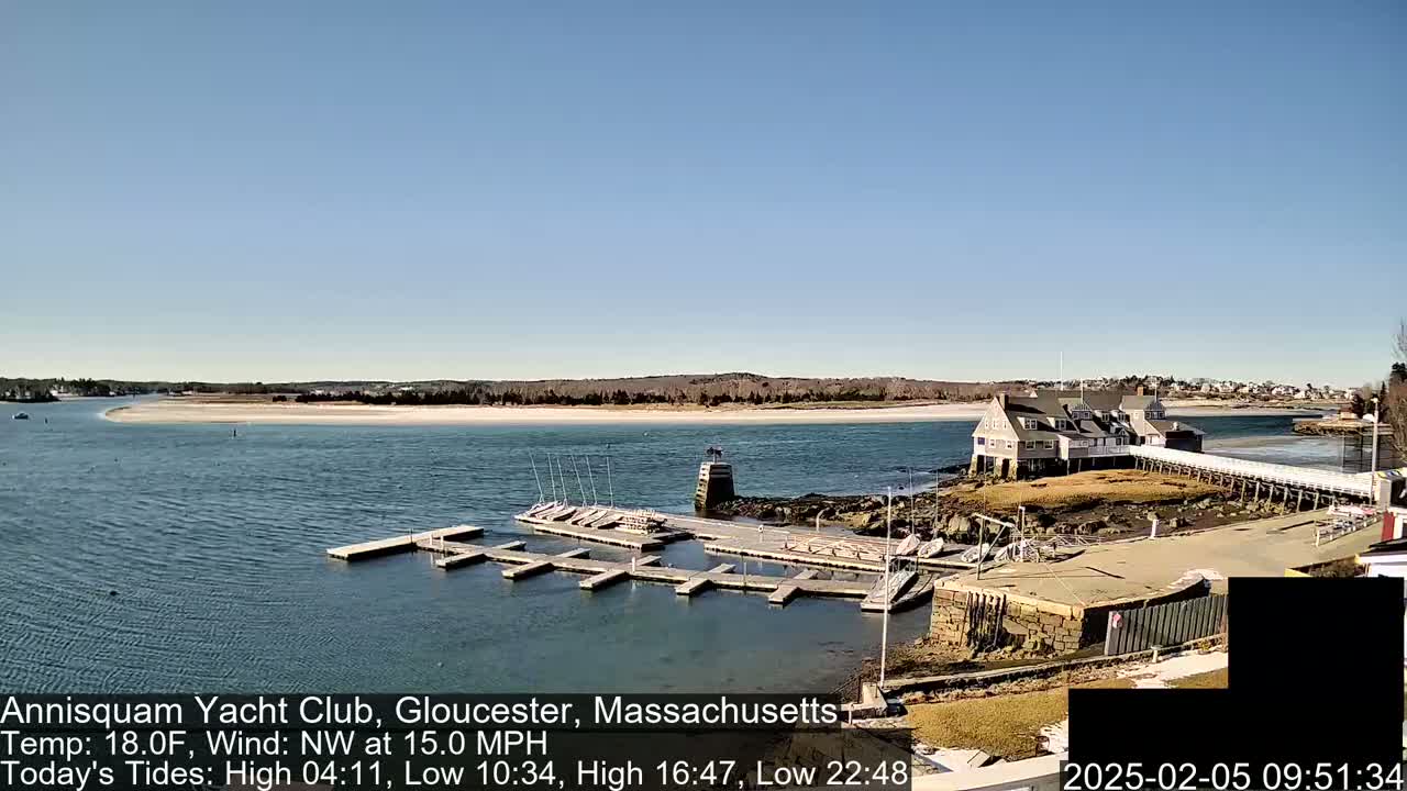 A sunny day reveals a marina with numerous small boats docked, situated next to a large clubhouse on a small peninsula extending into a calm body of water with a sandy beach in the background.