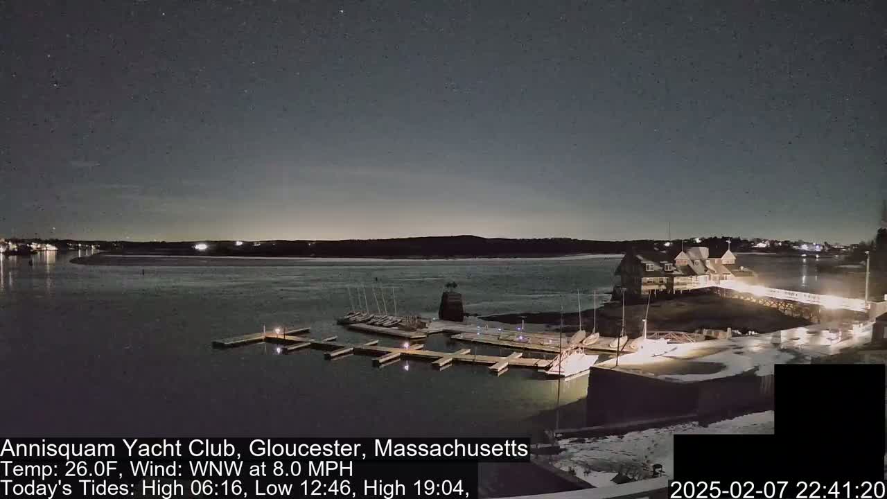 A nighttime view of a marina with docked sailboats, a clubhouse, and a partially snow-covered shoreline under a starry sky and a 26°F temperature with a west-northwest wind of 8.0 mph.