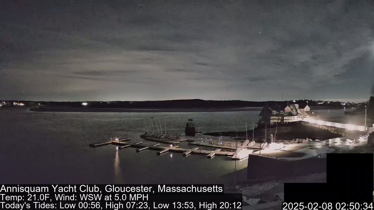 A nighttime view of a marina with boats docked, a clubhouse, and a calm body of water under a cloudy sky.