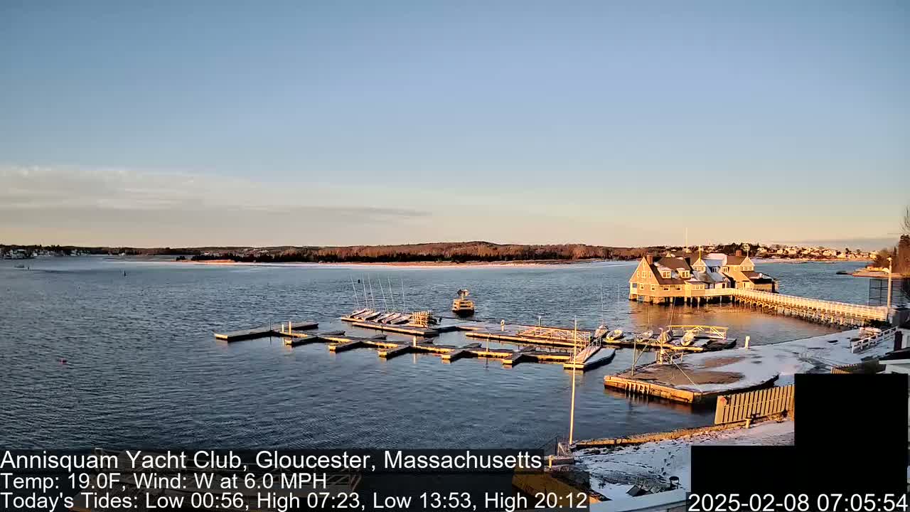 A calm body of water with a marina, a clubhouse, and docked sailboats under a clear, twilight sky with some snow on the ground.