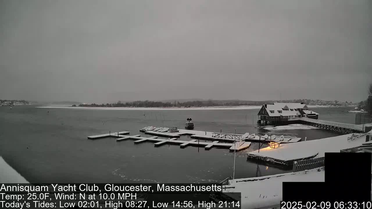 A snow-covered marina with a clubhouse and several docks sits on a calm, snowy body of water under an overcast sky.