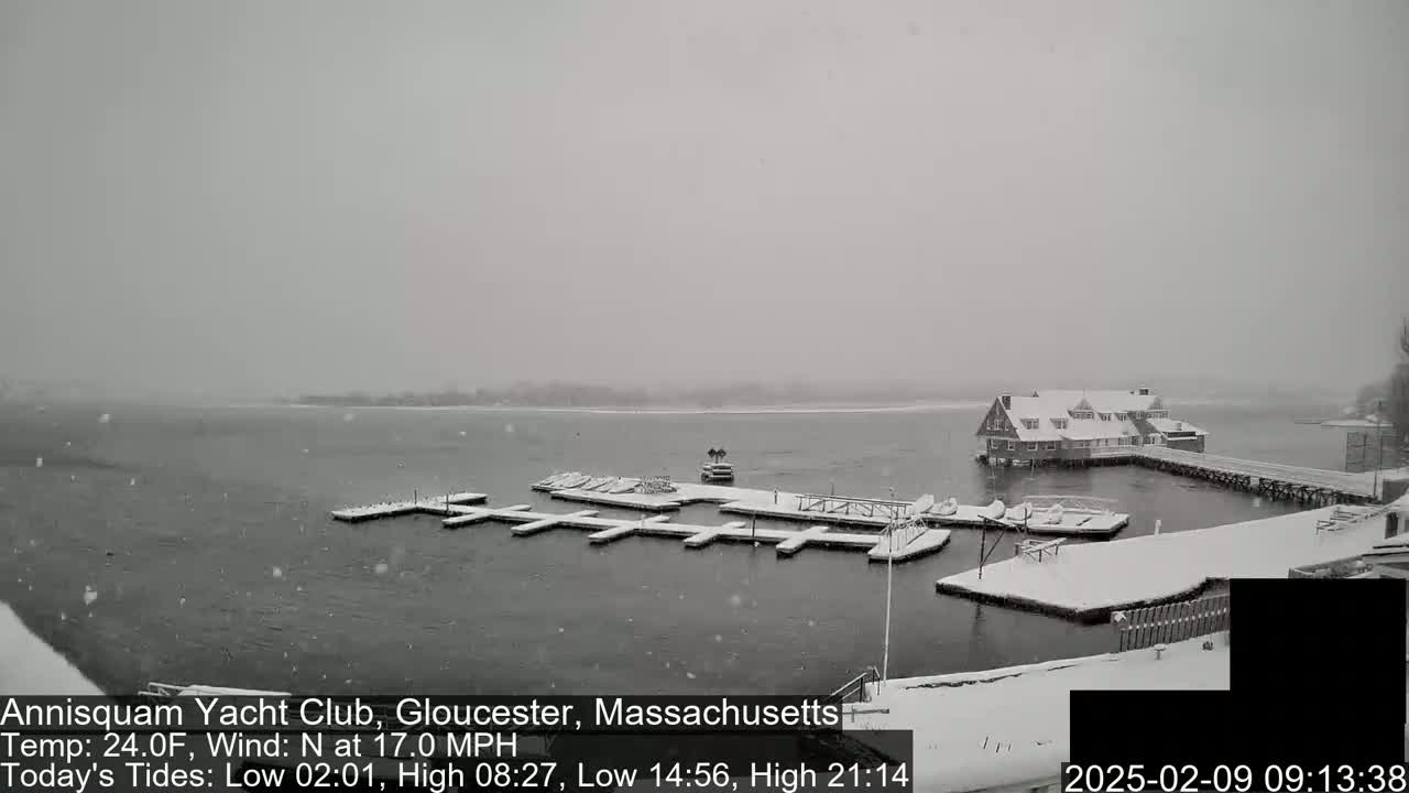 A snow-covered marina with boats docked and a building in the background during a snowfall.