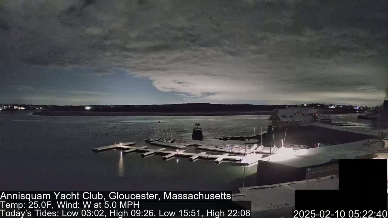 A nighttime view of a marina with boats docked, under a mostly cloudy sky, with a slight wind.