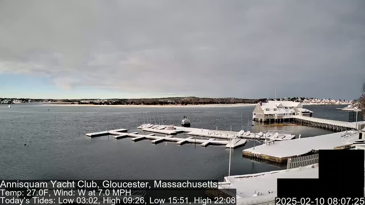 A snow-covered marina with sailboats docked, a clubhouse, and a partly cloudy sky.