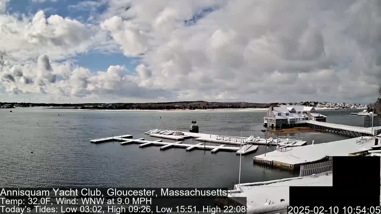 A snow-covered marina with boats docked at various slips, a white building, and a pier extends into a calm body of water under a partly cloudy sky.