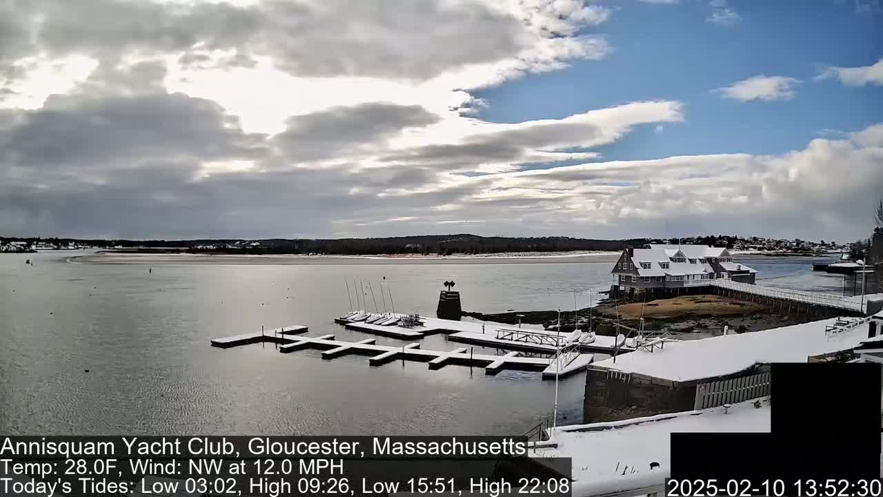 A snow-covered marina with boats docked and a building in the background under a partly cloudy sky.