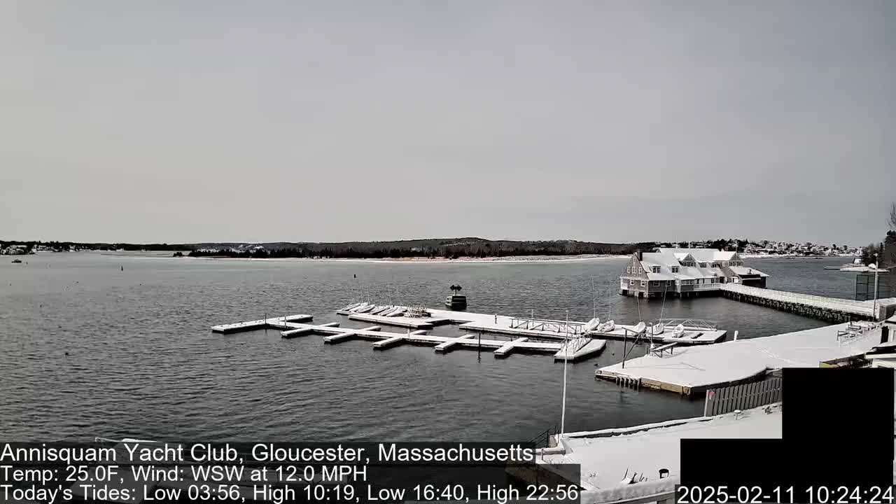 A snow-covered yacht club with a dock filled with boats sits on a body of water under a cloudy sky.