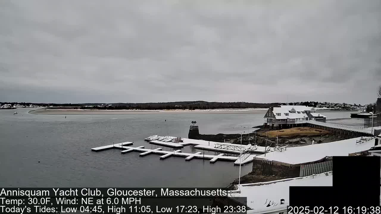 A snow-covered yacht club and marina sit on a calm, gray waterway under an overcast sky.