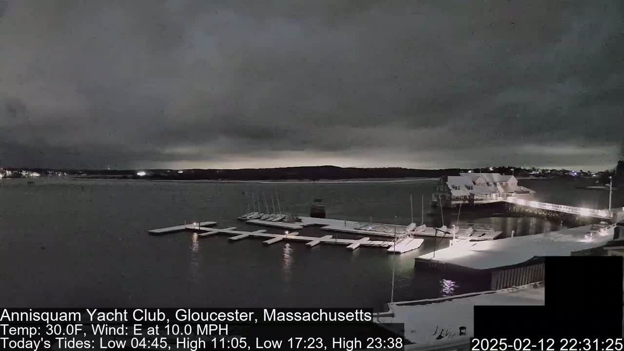 A snow-covered marina at night under a dark, cloudy sky, with boats docked and lights illuminating the scene.