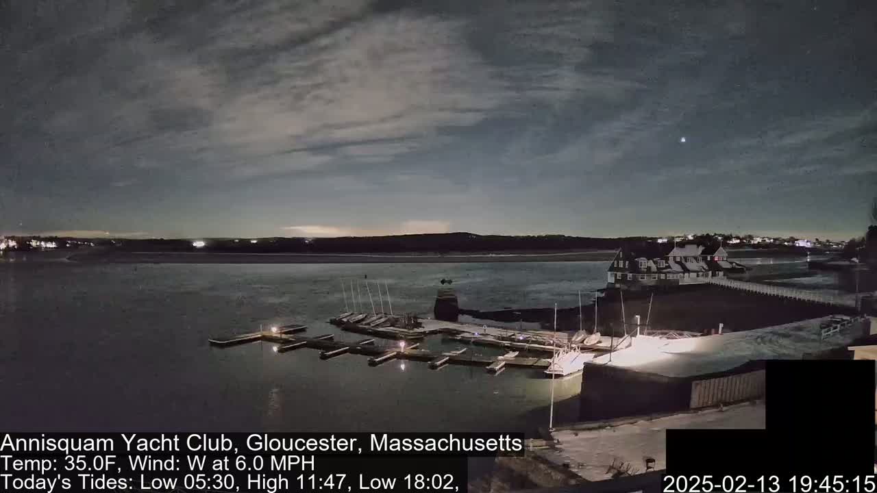 A nighttime view of a marina with docked sailboats under a partly cloudy sky, with a calm body of water and a building in the background.