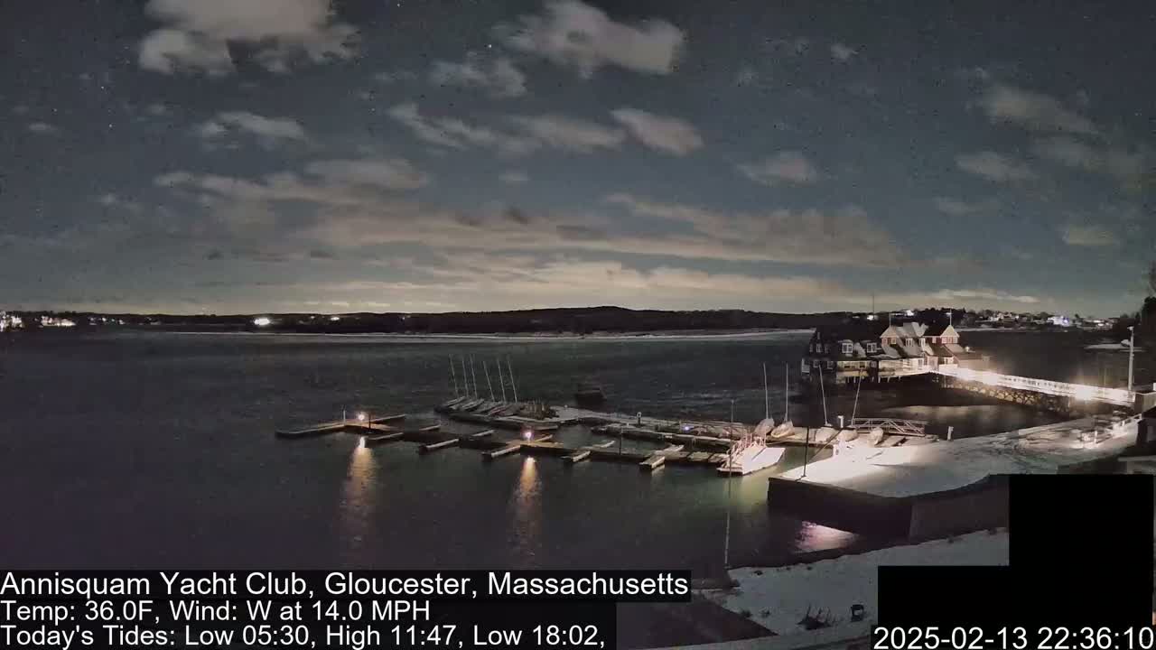A nighttime view of a marina with boats docked, under a partly cloudy sky with a 14 mph westerly wind.