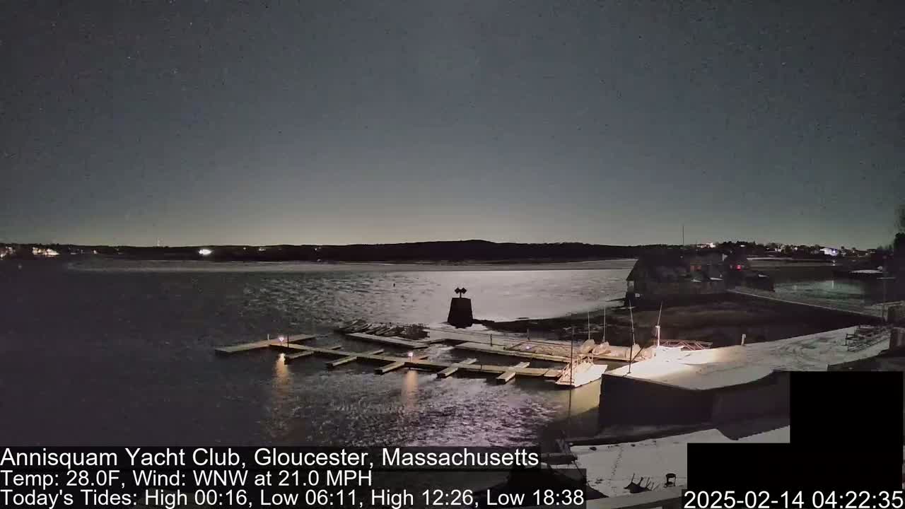 A nighttime view of a marina with docks and a small building, under a mostly clear sky with a WNW wind at 21 mph.