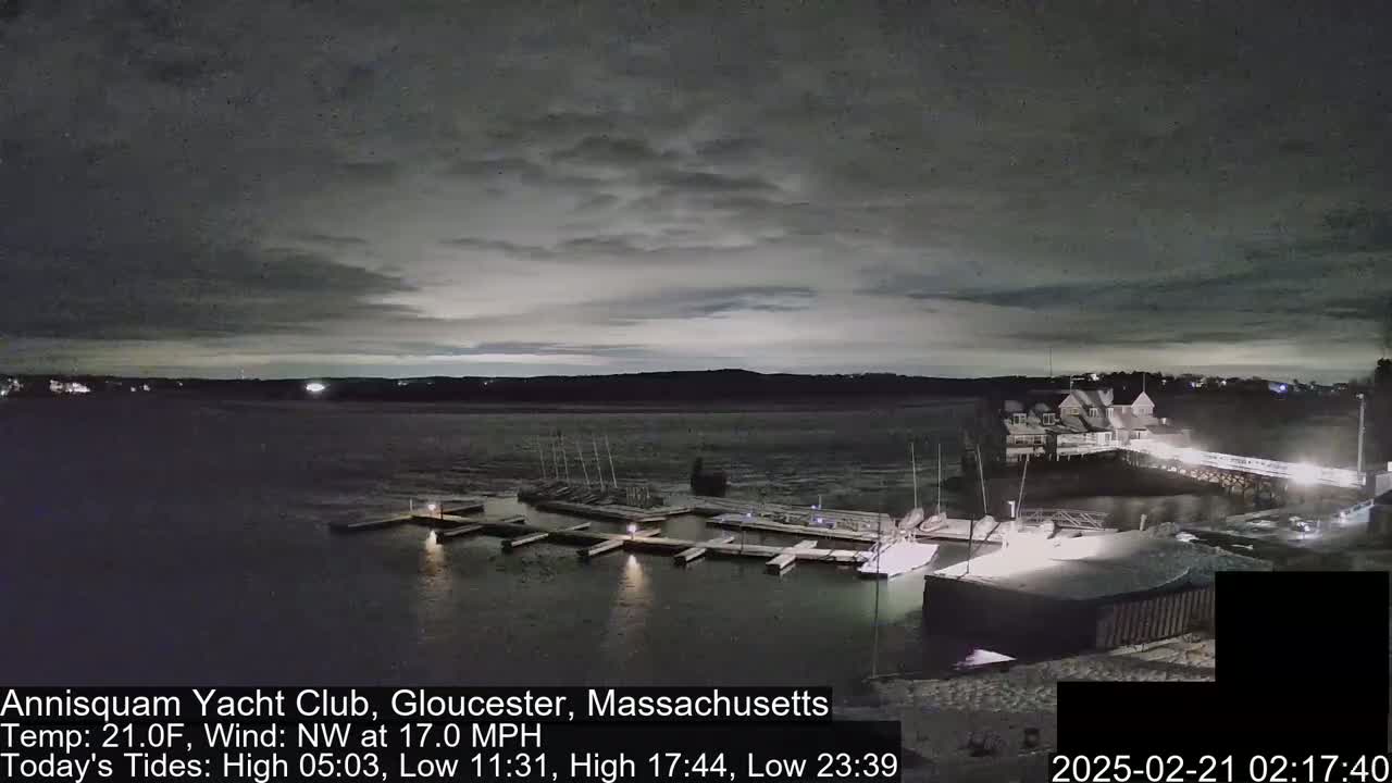 A nighttime view of a marina with boats docked, under a mostly cloudy sky, with a northwest wind at 17 mph and a temperature of 21 degrees Fahrenheit.