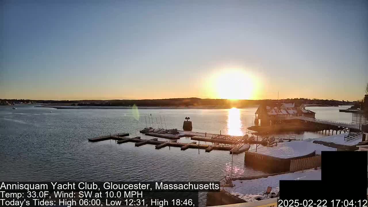A calm, sunlit waterfront scene at sunset shows a marina with boats and docks partially covered in snow.