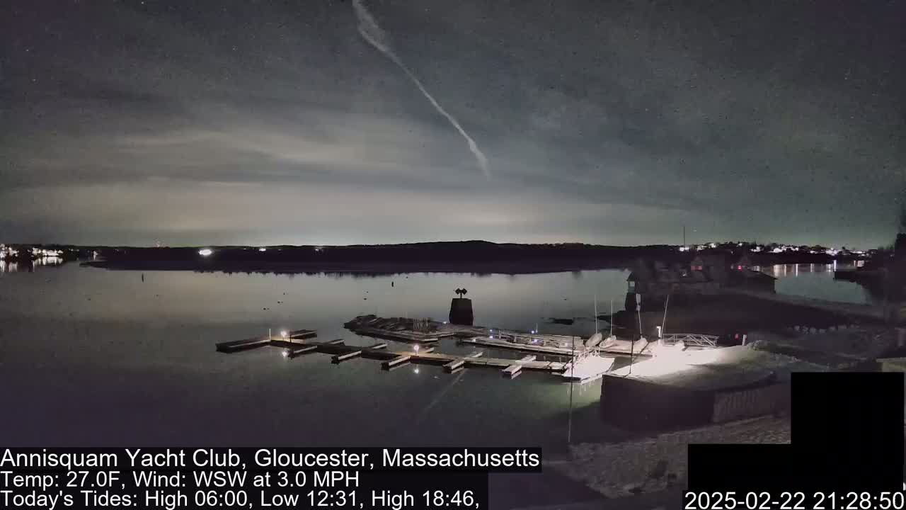 A nighttime view of a marina on a calm body of water under a partly cloudy sky with a faint contrail visible.