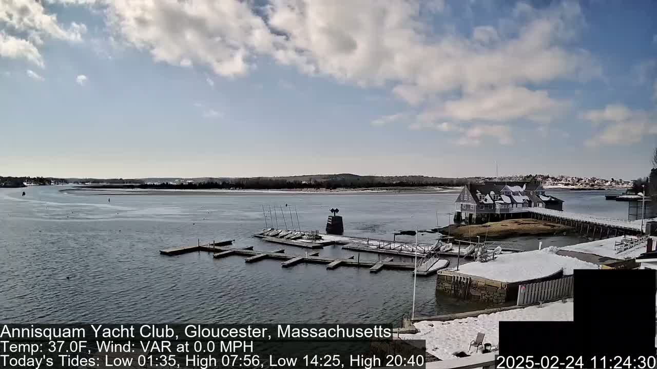 A partly cloudy day shows a snow-dusted yacht club and marina on a calm body of water.