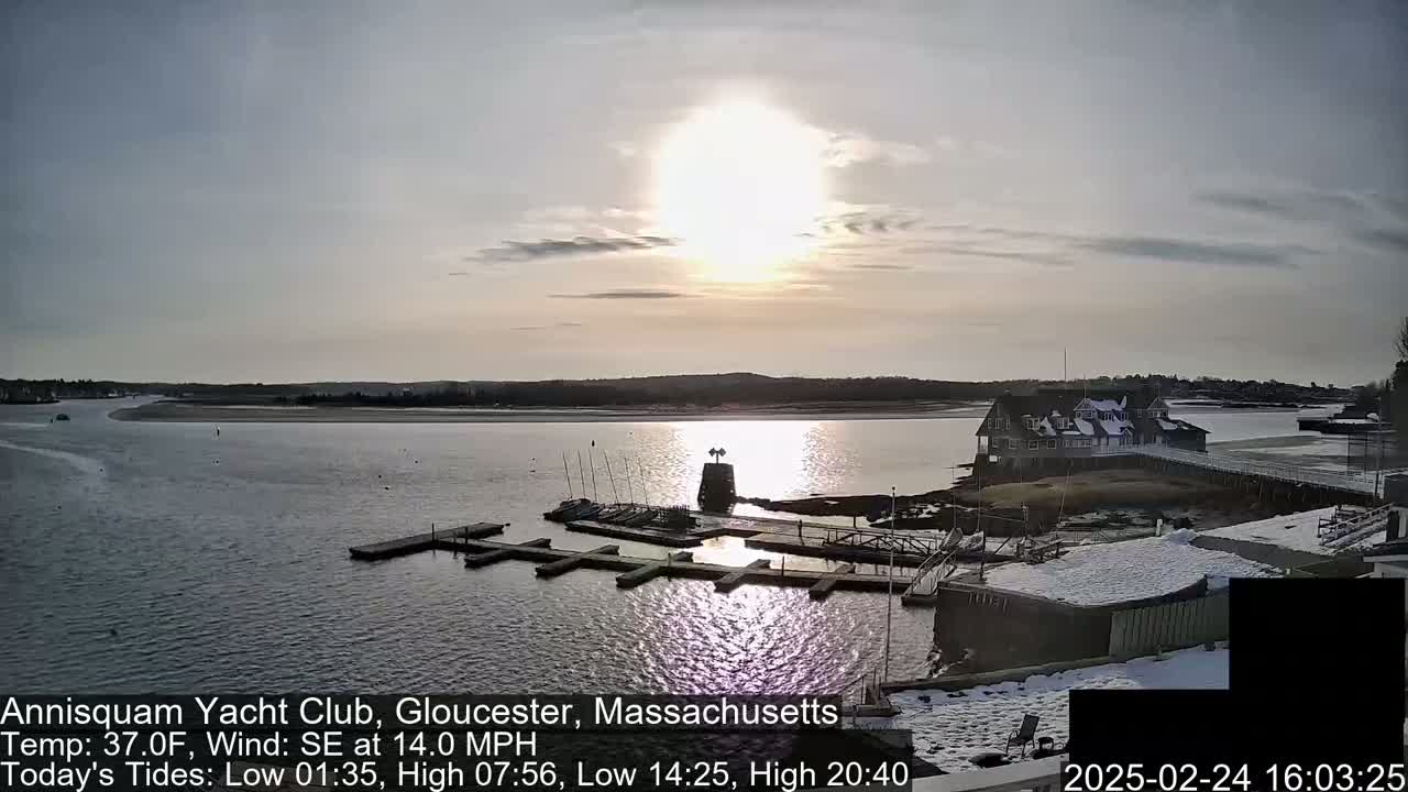 A marina with sailboats and a clubhouse is partially covered in snow, under a bright, mostly clear sunset sky.