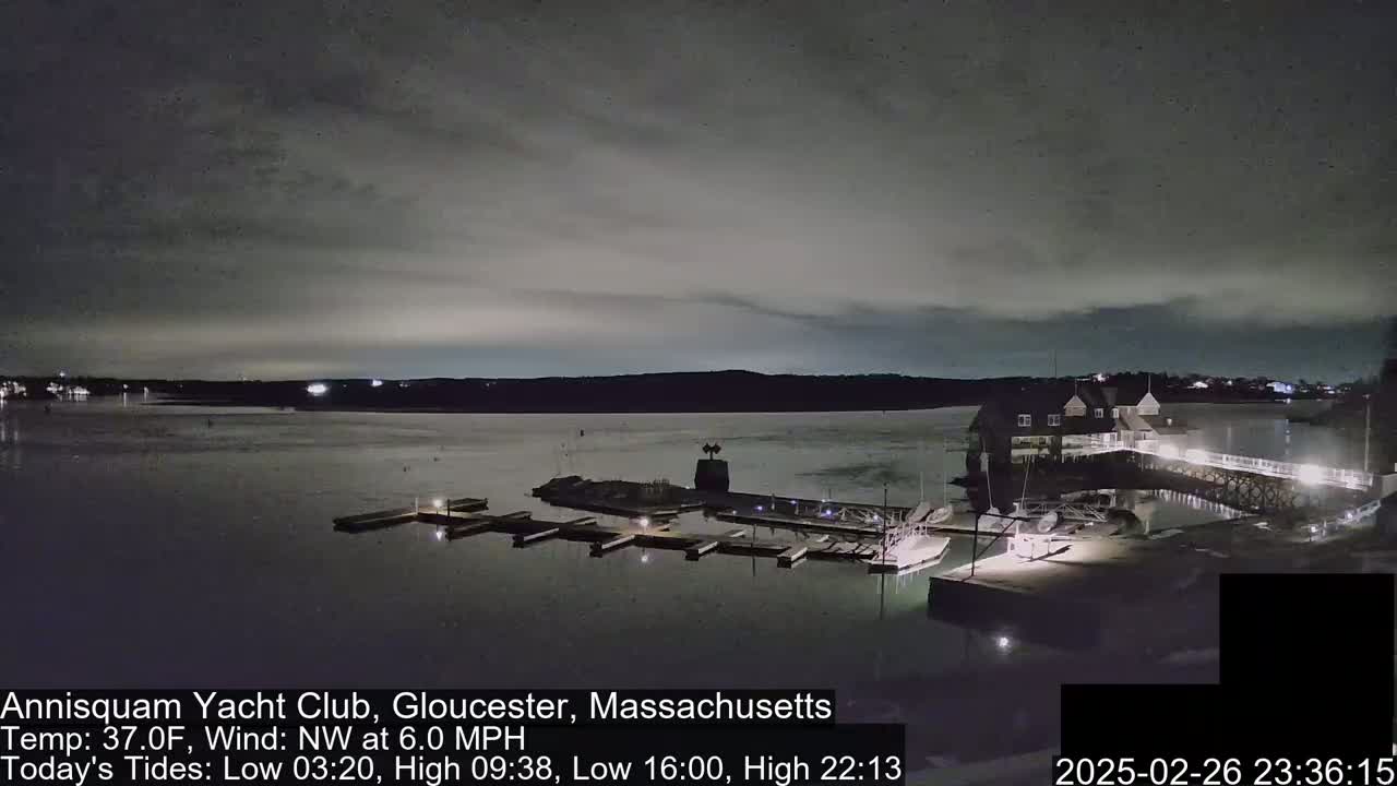 A dark nighttime view of a calm body of water with a marina and a clubhouse under a cloudy sky.