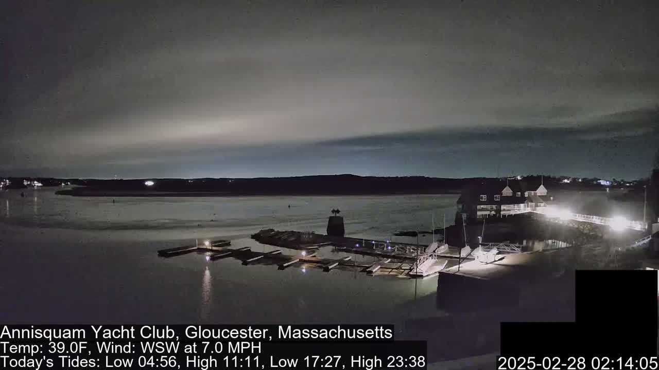 A nighttime view of a calm harbor with docked boats and a building under a mostly cloudy sky.