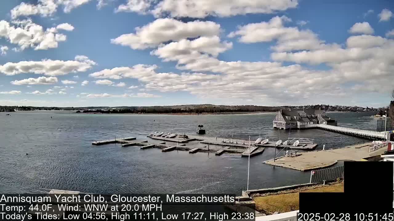 A partly cloudy day shows a marina with numerous boats docked, a clubhouse, and a long pier extending into the water under a partly cloudy sky.