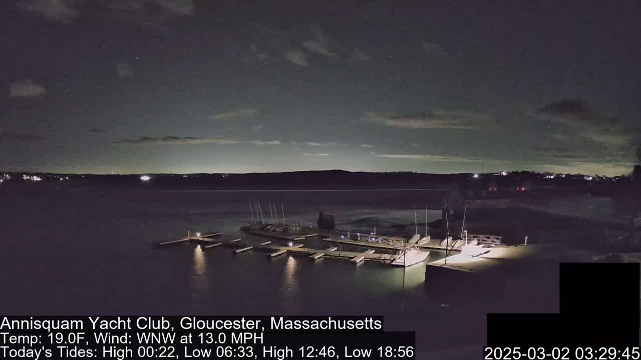 A nighttime view of a marina with boats docked at piers under a partly cloudy sky with a 13 mph wind.