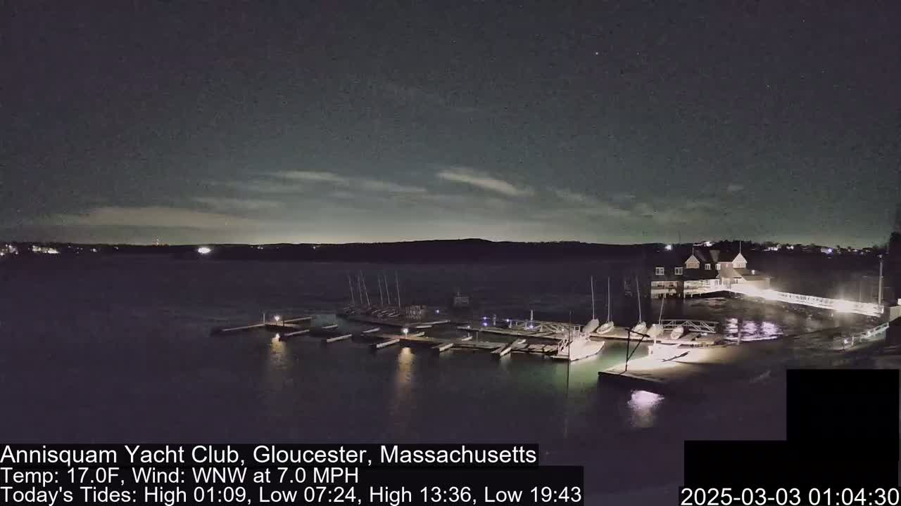 A nighttime view of a marina with boats docked, under a partly cloudy sky and a 7 mph wind.