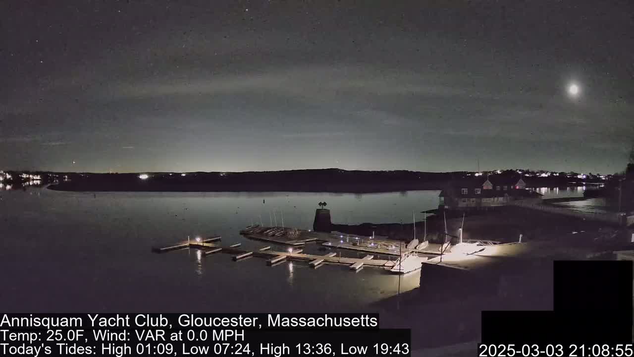 A nighttime view of a calm harbor with a marina, a building, and a bright moon in the sky.