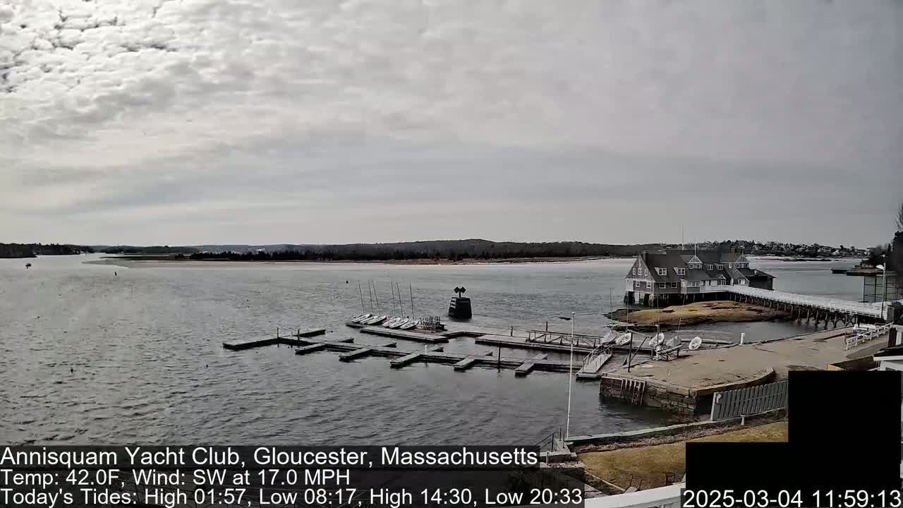 A calm body of water with a yacht club and several small sailboats docked, under a cloudy sky.