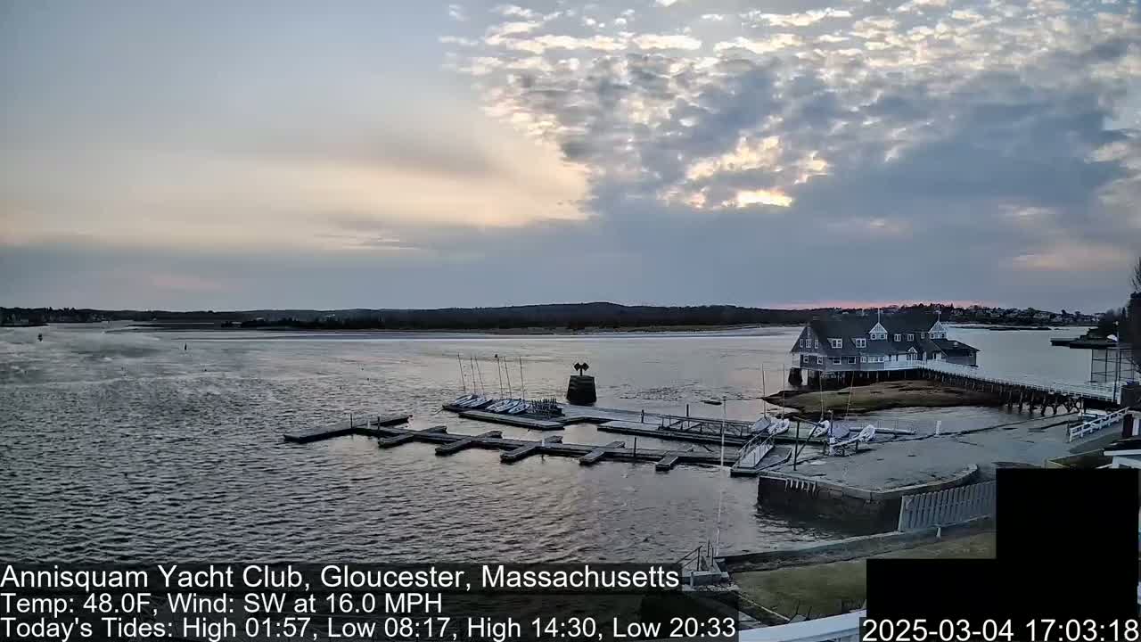 A calm body of water with docked sailboats and a clubhouse under a partly cloudy sky.