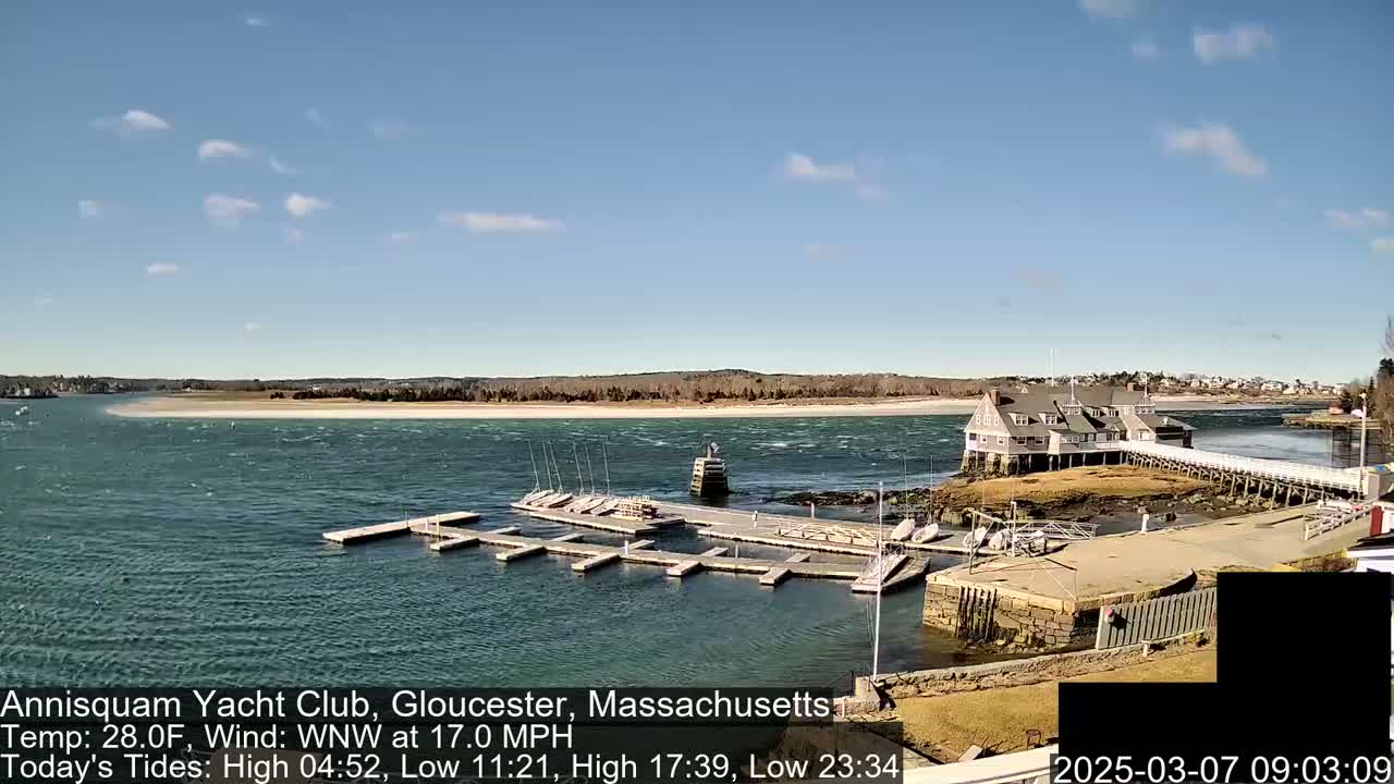 A yacht club sits on a small island connected to the mainland by a pier, with boats moored at the docks in a choppy body of water under a mostly sunny sky.