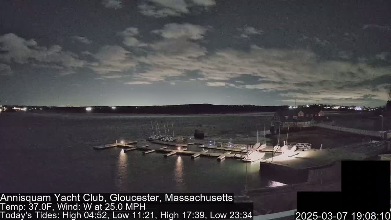 A nighttime view of a marina with boats docked, under a partly cloudy sky, and a strong west wind.