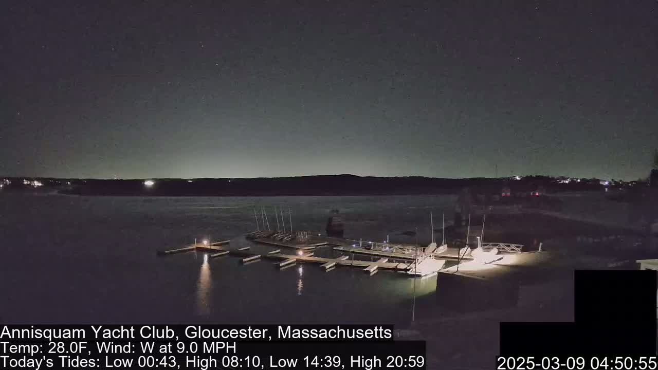 A nighttime view of a marina with boats docked, under a partly cloudy sky and a 9.0 mph wind.