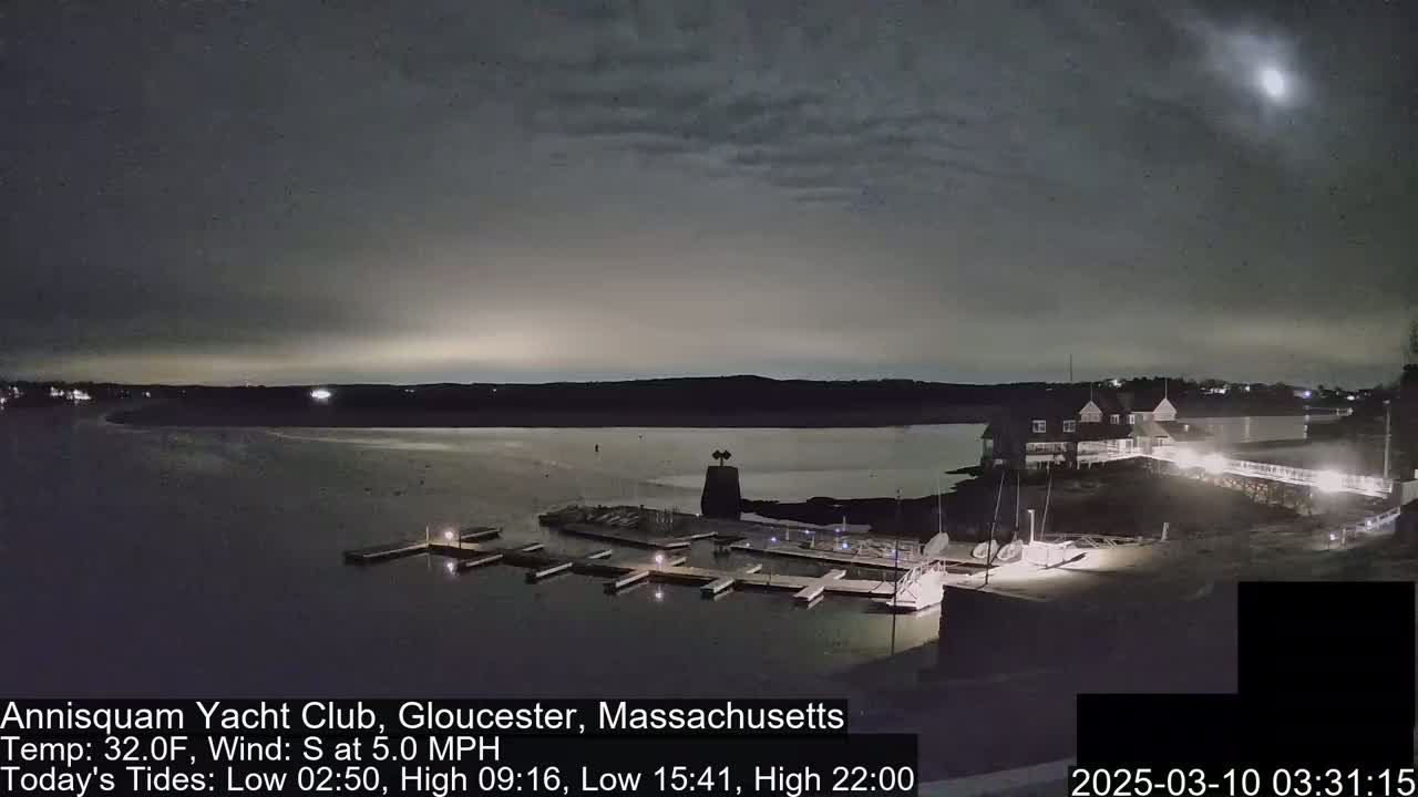 A nighttime view of a marina with docked boats, a clubhouse, and calm water under a mostly cloudy sky.
