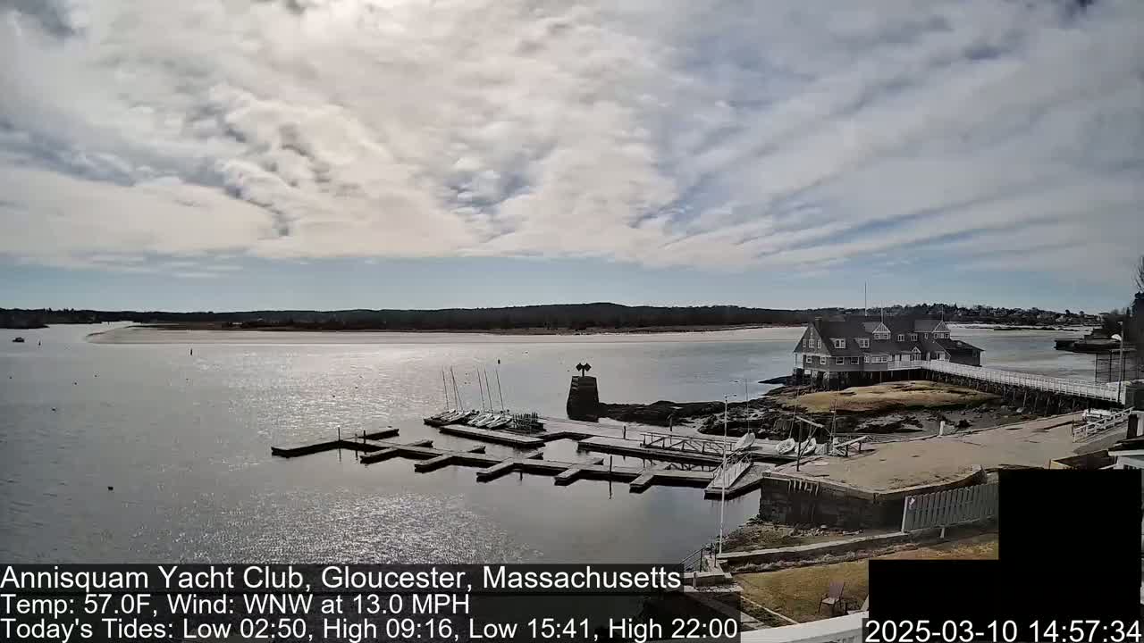 A partly cloudy day shows a yacht club on a small island connected to the mainland by a pier, with sailboats docked in the calm water.