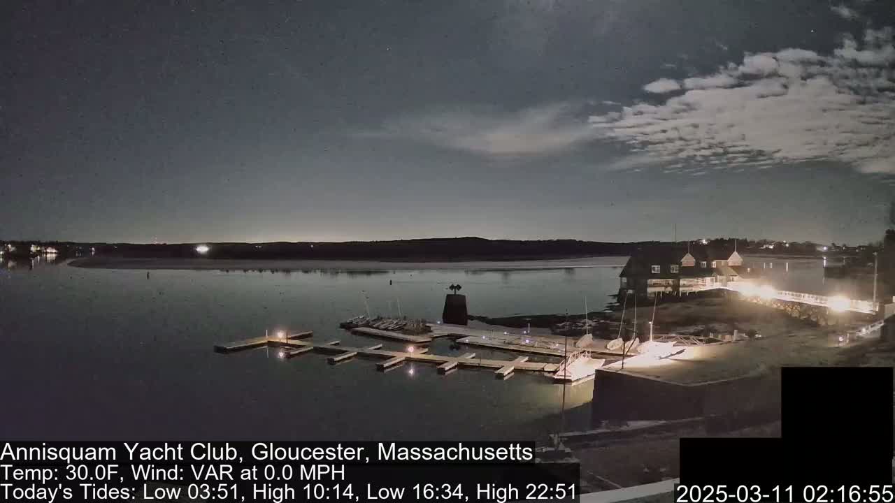 A nighttime view of a marina with boats docked, a clubhouse, and a calm body of water under a partly cloudy sky.