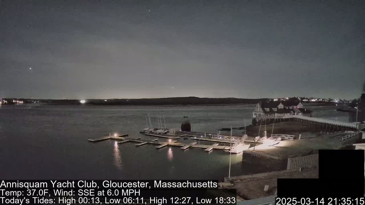 A nighttime view of a marina with boats docked, lit by artificial lights under a dark, mostly clear sky.
