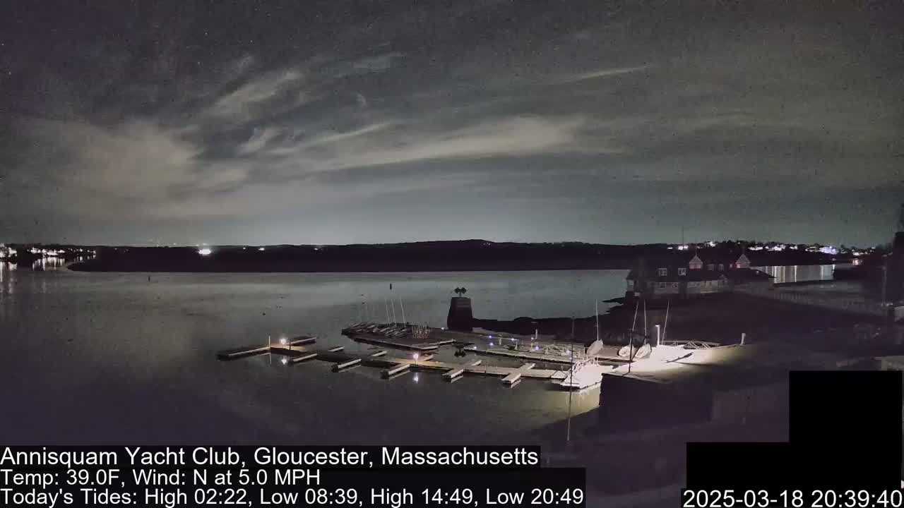 A nighttime view of a marina with docked boats, a clubhouse, and a calm body of water under a partly cloudy sky.